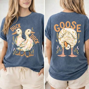 May include: A blue t-shirt with a duck and goose design. The front displays two ducks with the word "DUCK" above each. The back shows a goose bending over with the word "GOOSE" above it. The shirt has rolled-up sleeves.