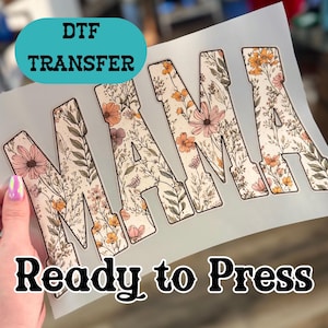 May include: A DTF transfer sheet with the word "MAMA" in large, block letters. The letters are filled with a floral pattern in shades of pink, orange, and cream. The text "Ready to Press" is below the word "MAMA". A teal oval shape contains the text "DTF TRANSFER".