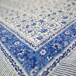 May include: A white and blue floral patterned textile. The design features a border of blue floral motifs and stripes. The fabric has a soft, faded appearance, suggesting a vintage or handcrafted style. The overall aesthetic is reminiscent of traditional block printing.