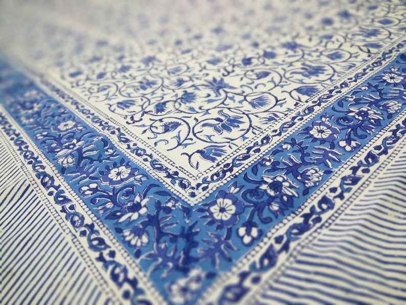 May include: A white and blue floral patterned textile. The design features a border of blue floral motifs and stripes. The fabric has a soft, faded appearance, suggesting a vintage or handcrafted style. The overall aesthetic is reminiscent of traditional block printing.