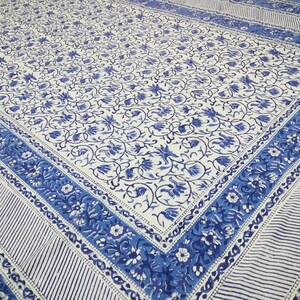 May include: A white and blue floral patterned textile. The design features a repeating pattern of blue flowers and vines on a white background. The border has a striped pattern and floral design. The textile is likely a tablecloth or bedspread.