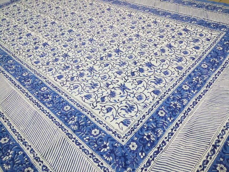 May include: A white and blue floral patterned textile. The design features a repeating pattern of blue flowers and vines on a white background. The border has a striped pattern and floral design. The textile is likely a tablecloth or bedspread.