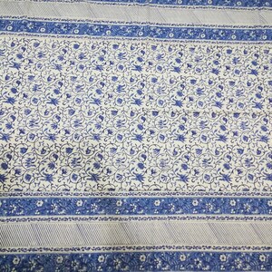 May include: A white and blue floral patterned textile. The design features a central field of blue floral motifs on a white background, framed by a blue border with floral and striped patterns. The textile is likely a tablecloth or bedspread.