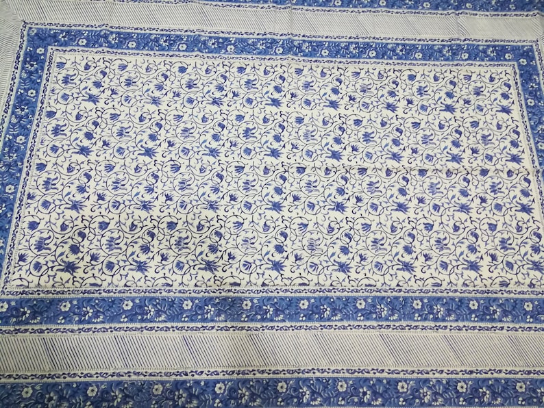 May include: A white and blue floral patterned textile. The design features a central field of blue floral motifs on a white background, framed by a blue border with floral and striped patterns. The textile is likely a tablecloth or bedspread.