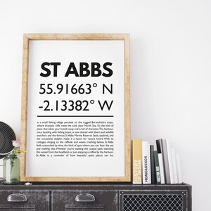May include: A framed print with the text "ST ABBS" and coordinates 55.91663° N, -2.13382° W. The print is in a wooden frame and rests on a dark metal cabinet with books and a vase. The text describes a fishing village.