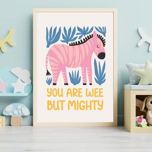 May include: A framed wall art print featuring a pink zebra with dark stripes standing among blue stylised leaves. Below the zebra, the text reads "YOU ARE WEE BUT MIGHTY" in yellow capital letters. The print is set against a cream background.