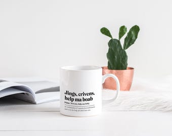 May include: White ceramic mug with the text "Jings, crivens, help ma boab" and its definition. A green cactus in a copper-colored pot sits behind the mug. An open book is on the left.