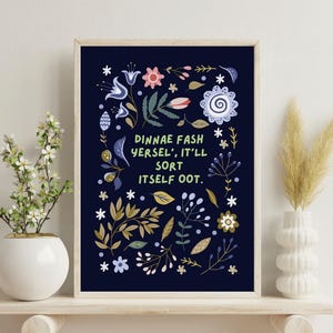 May include: A framed art print with a navy blue background, decorated with floral illustrations in white, gold, pink, and blue. The print includes the text "DINNAE FASH YERSEL', IT'LL SORT ITSELF OOT."