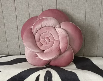 Puede incluir: Un cojín en forma de rosa, de color rosa empolvado, descansa sobre una manta con estampado de cebra. El cojín imita una rosa, con pétalos superpuestos y un centro en espiral. El fondo presenta un cabecero gris con líneas verticales.