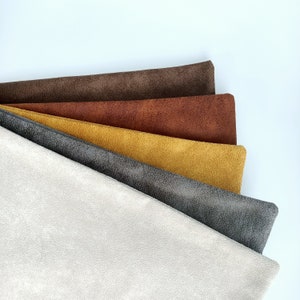 May include: A selection of fabric swatches in shades of cream, grey, mustard yellow, rust, and brown. The swatches are fanned out, displaying the different textures and colours of the material. The fabric has a suede-like texture. Each swatch is approximately 30 cm long.