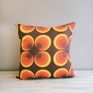 Puede incluir: Una almohada marrón con un patrón geométrico de círculos naranjas y amarillos. Los círculos están dispuestos en un patrón repetitivo.