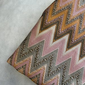 Op de afbeelding: Een textielstof met een herhalend chevronpatroon in tinten oudroze, goudgeel en metallic zilvergrijs. Het materiaal heeft een getextureerd, geweven uiterlijk met duidelijke zigzaglijnen die diagonaal over het oppervlak lopen.