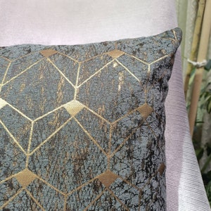 May include: A square cushion with a geometric gold pattern on a textured grey background. The cushion has a soft appearance, ideal for adding elegance to any room. The design features intersecting lines and diamond shapes. The cushion is approximately 40cm x 40cm.