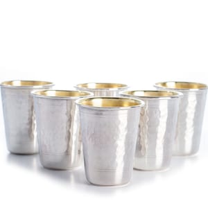 Pode incluir: Seis copos de prata com bordas douradas. Os copos estão dispostos em fila sobre um fundo branco.