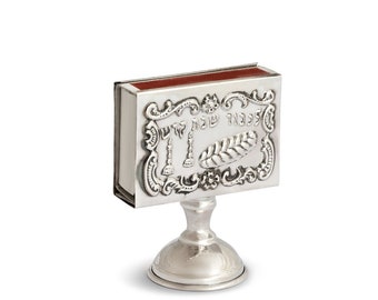 May include: Silver matchbox holder with an ornate design and the Hebrew words "Shabbat Shalom" engraved on the front. The holder has a small base.