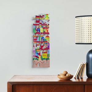 Pode incluir: Uma peça de arte de parede de madeira colorida com letras hebraicas e um design de cidade. As letras são dispostas de uma forma que cria a imagem de edifícios e um céu com nuvens. A peça é pintada em cores brilhantes, incluindo vermelho, amarelo, azul, verde e roxo.