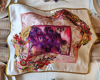May include: A white embroidered cushion with a gold trim and a pink and purple floral design. The cushion features a central square with a Hebrew inscription.