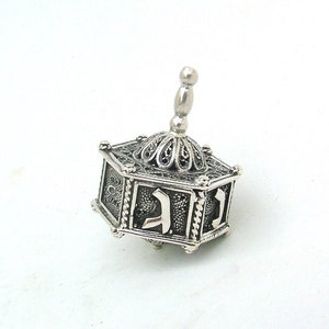 Puede incluir: Un trompo de plata con intrincados diseños de filigrana y letras hebreas en cada lado. El trompo es un juguete tradicional judío que se utiliza durante Janucá.