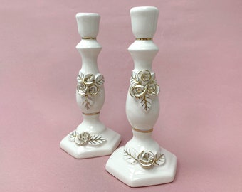 May include: Two white ceramic candle holders with gold accents and floral designs. Each holder features a cluster of three white roses with gold centers and green leaves.