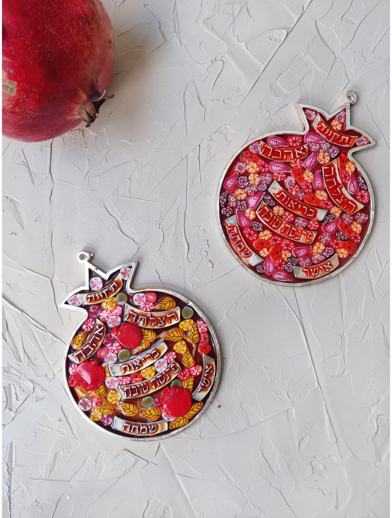K&ouml;nnte beinhalten: Zwei silberne Granatapfel-Ornamente mit roten, gelben und wei&szlig;en Mosaikmustern. Die Ornamente zeigen hebr&auml;ischen Text und sind ein traditionelles Symbol von Rosh Hashanah.