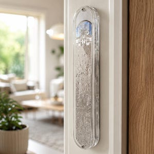 May include: A clear, rectangular mezuzah case with a detailed silver-colored relief design of a cityscape. The case is mounted on a white door frame, with a glimpse of a living room in the background. The design includes architectural elements.