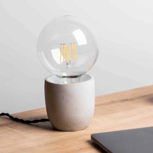 Op de afbeelding: Een witte betonnen tafellamp met een heldere glazen bol als lamp. De lamp staat op een houten oppervlak.
