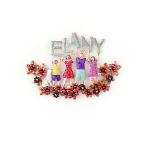 May include: A handcrafted metal family sculpture with the name "ELANY" held aloft by figures. The figures are dressed in various colors and stand above a floral arrangement with red and gold accents. A white dog is also present.