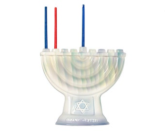 May include: White ceramic menorah with blue and red candles. The menorah has a Star of David design and the Hebrew words "Happy Hanukkah" on the base.