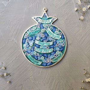 K&ouml;nnte beinhalten: Ein silberner Granatapfel-Anh&auml;nger mit blauem und gr&uuml;nem Emaille und hebr&auml;ischem Text. Der Anh&auml;nger ist mit kleinen blauen Blumen und Bl&auml;ttern verziert.