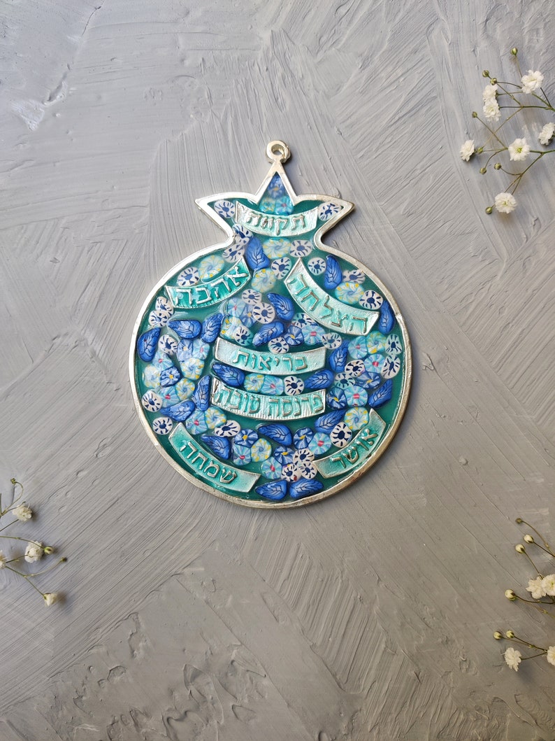 K&ouml;nnte beinhalten: Ein silberner Granatapfel-Anh&auml;nger mit blauem und gr&uuml;nem Emaille und hebr&auml;ischem Text. Der Anh&auml;nger ist mit kleinen blauen Blumen und Bl&auml;ttern verziert.