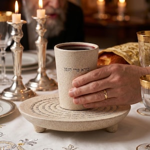 May include: A beige ceramic cup with dark liquid, held on a matching round plate with Hebrew text. The cup has Hebrew text on it. Silver candle holders and lit candles are in the background, along with a person.