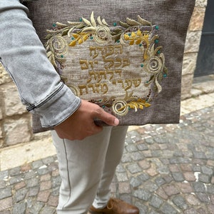 Puede incluir: Una bolsa de tela marr&oacute;n con dise&ntilde;os florales bordados y texto en hebreo. La bolsa la lleva una persona que lleva una chaqueta vaquera gris claro y pantalones beige.