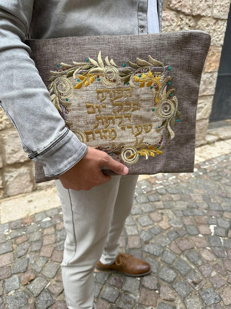Puede incluir: Una bolsa de tela marr&oacute;n con dise&ntilde;os florales bordados y texto en hebreo. La bolsa la lleva una persona que lleva una chaqueta vaquera gris claro y pantalones beige.