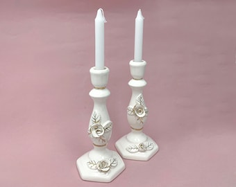 May include: Two white ceramic candle holders with gold accents and white roses. The candle holders have a decorative design with leaves and flowers.