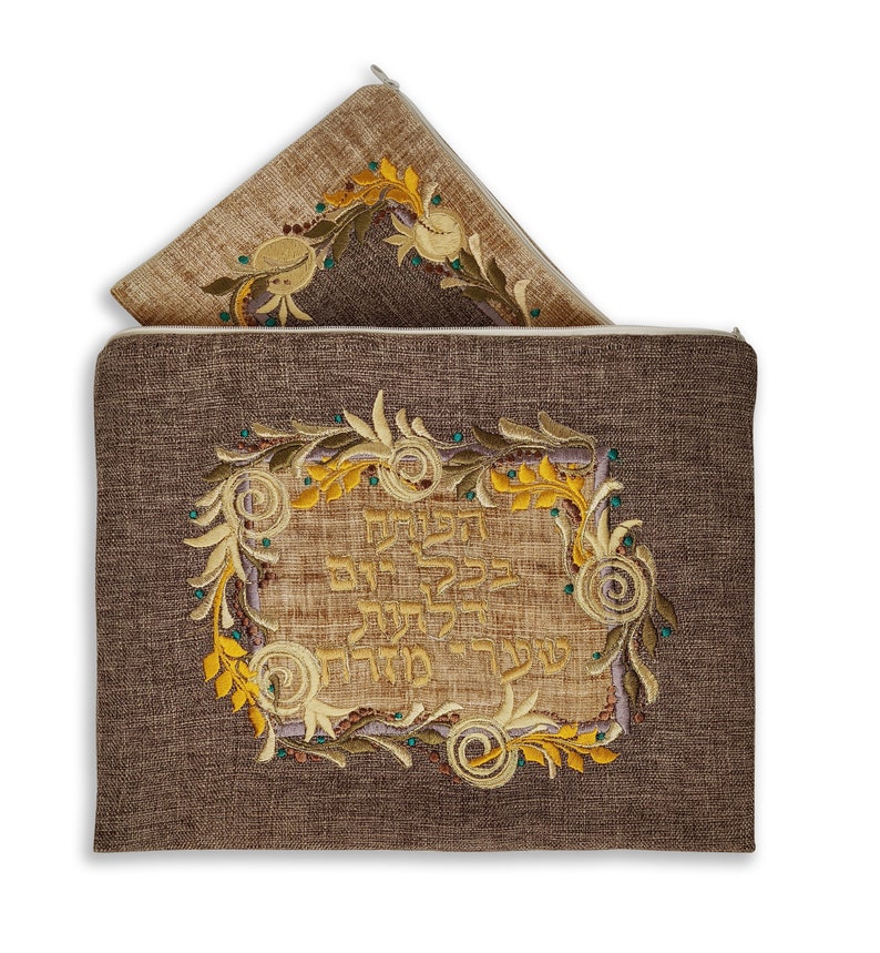 Puede incluir: Una cubierta de challah de lino marr&oacute;n con cierre de cremallera. La cubierta presenta un dise&ntilde;o bordado de una corona floral con texto hebreo en el centro. El texto dice "Bendito seas t&uacute;, Se&ntilde;or nuestro Dios, Rey del universo, que creas el fruto de la vid."