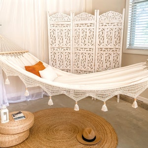 May include: A white hammock with tassels hangs in a room with a white screen and a brown rug. A straw hat sits on the rug.