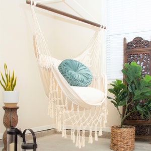 May include: A cream-colored macrame hammock chair with a wooden bar, hanging from the ceiling. A teal round pillow rests inside the chair. Decorative lanterns, a potted plant, and a patterned rug complete the cozy, bohemian-style scene.