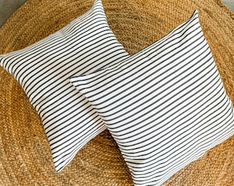 May include: Two square throw pillows with black and white horizontal stripes. The pillows are placed on a round, woven jute rug. The pillows have a simple, modern design, suitable for a living room or bedroom.