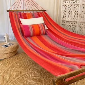 May include: A fabric hammock featuring a vibrant striped pattern in shades of red, orange, and pink, suspended by a dark wooden spreader bar. The hammock includes a rectangular throw pillow with matching stripes and a solid white rectangular pillow.