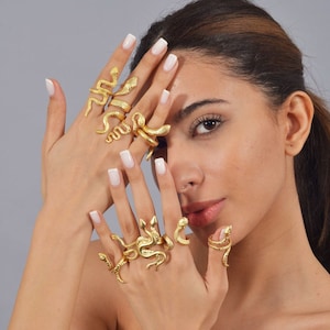 May include: A woman's hand wearing multiple gold snake rings. The rings are stacked on each finger and are a statement piece of jewelry.