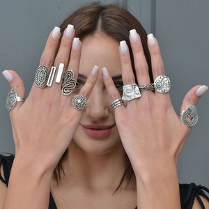 Può includere: Le mani di una donna con una varietà di anelli d'argento con disegni intricati. Gli anelli sono impilati su più dita e presentano motivi geometrici, spirali e una forma di cuore.