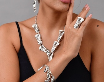 May include: A silver necklace and bracelet set featuring a repeating design of stylized faces. The necklace is a v-shaped chain with the face pendants hanging from it. The bracelet is a chain with several face pendants dangling from it.