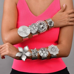 Può includere: Un set di sei braccialetti in metallo argentato con diversi design, tra cui un fiore, una stella, un cerchio e una rosa. I braccialetti sono indossati sul polso di una persona che indossa una canotta rosa.