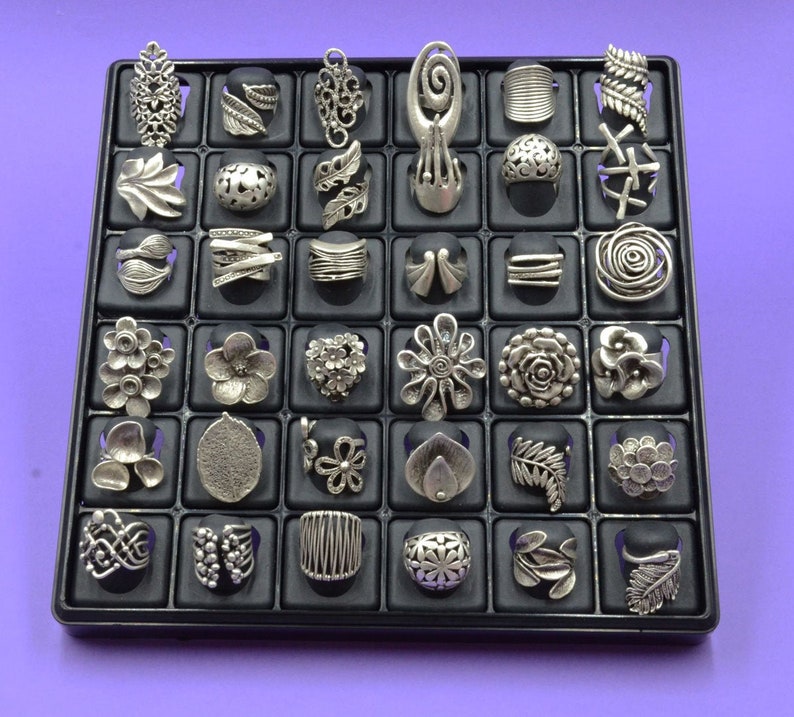 May include: A display case filled with silver rings. The rings are arranged in a grid pattern and feature a variety of designs, including floral, geometric, and abstract.