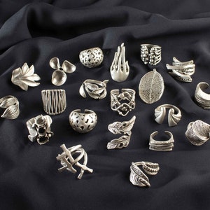 May include: A collection of 20 silver rings featuring a variety of designs, including leaves, feathers, and geometric patterns. The rings are displayed on a black fabric background.