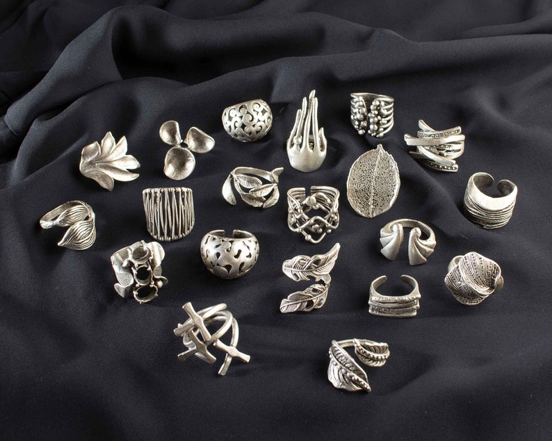 May include: A collection of 20 silver rings featuring a variety of designs, including leaves, feathers, and geometric patterns. The rings are displayed on a black fabric background.