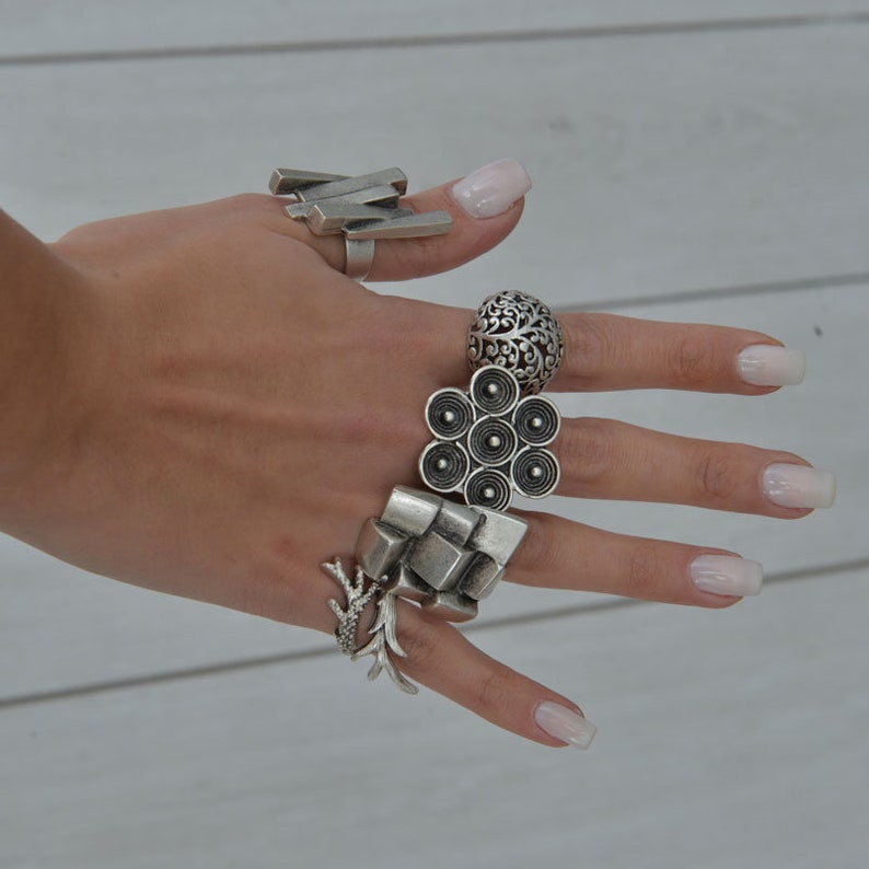 May include: A hand wearing five silver rings. The rings are all different designs, including a geometric design, a floral design, and a design with circles.