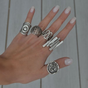 May include: A hand wearing five silver rings. The rings are all different styles, including a ring with a circle design, a ring with a lattice design, a ring with a wavy design, a ring with a stacked bar design, and a ring with a flower design.
