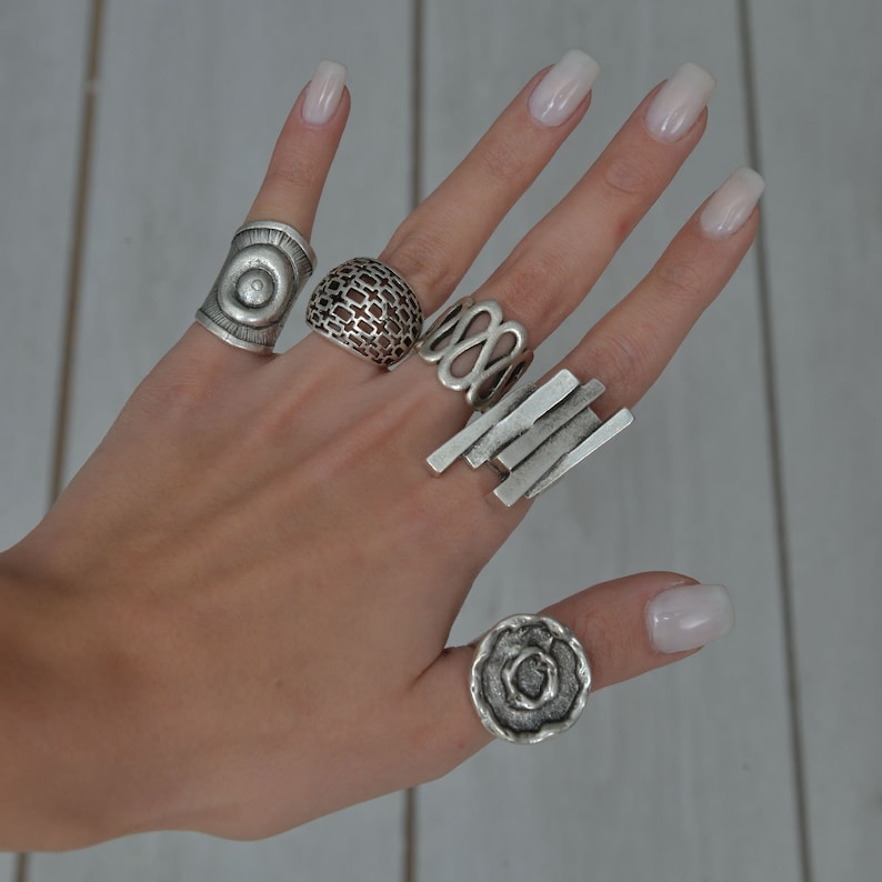 May include: A hand wearing five silver rings. The rings are all different styles, including a ring with a circle design, a ring with a lattice design, a ring with a wavy design, a ring with a stacked bar design, and a ring with a flower design.