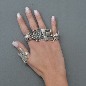 May include: A hand wearing five silver rings. The rings are all different styles, including a floral design, a geometric design, and a design with multiple circles.